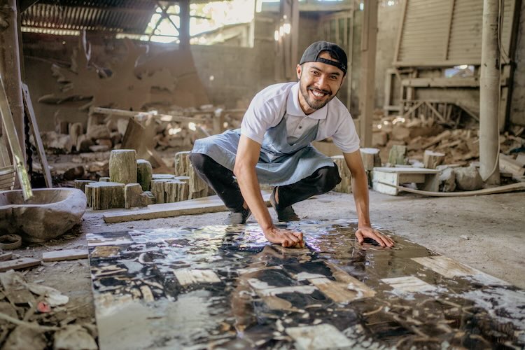 Smiling stonemason squats while washing large squares of stone in the workshop